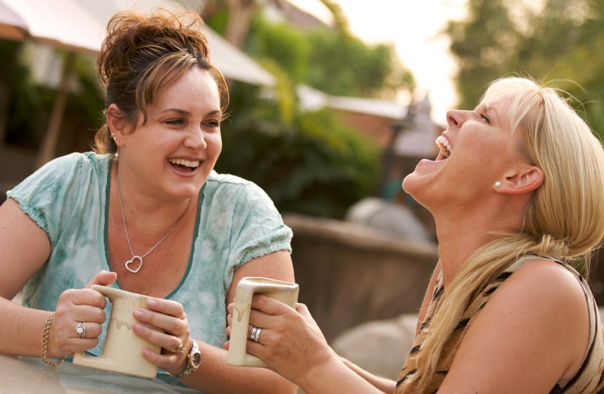 Two laughing women, each holding a cup, they are sitting outside