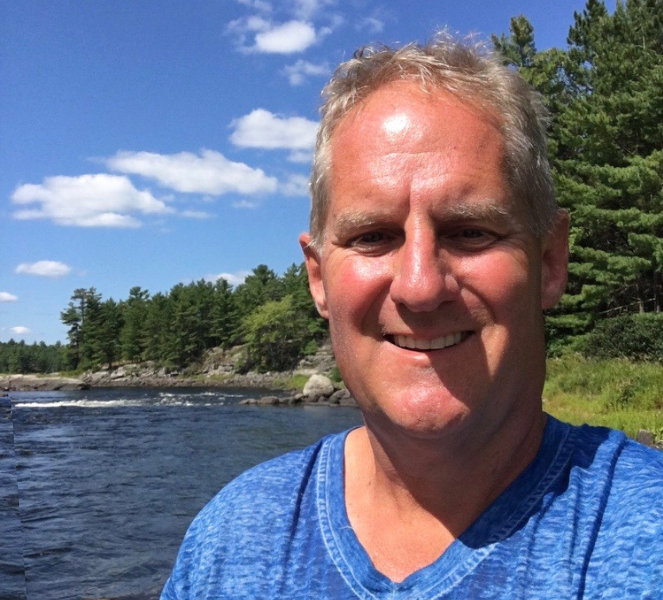 Keith Stevens in front of a beautiful lake surrounded by pine trees