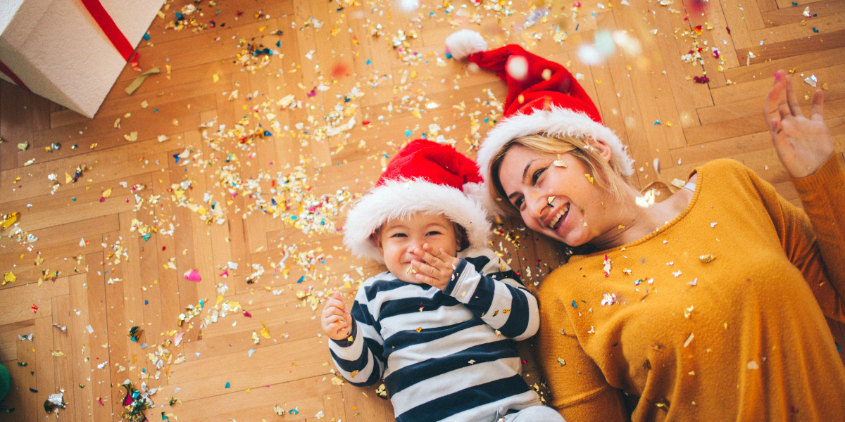 A mother and child laying on the floor wearing their santa hats and laughing. There's confetti scattered on the floor.