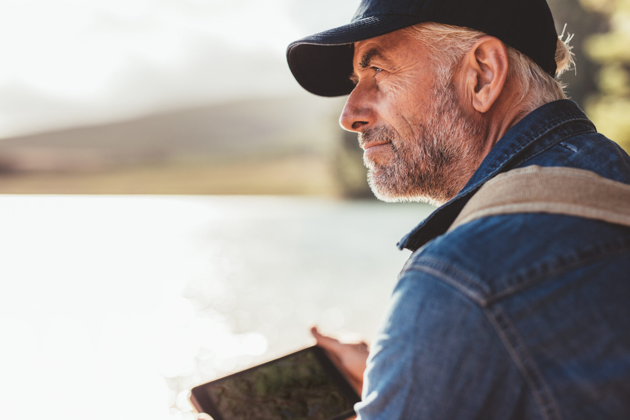 Close up portrait of mature man wearing cap sitting at a lake and looking at a view.